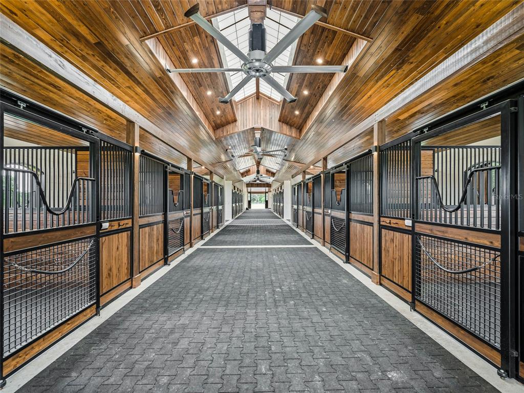 A long horse stable aisle features wood-paneled ceilings, black metal stalls, and a paved floor — 2380 NW 72ND COURT, OCALA, FL
