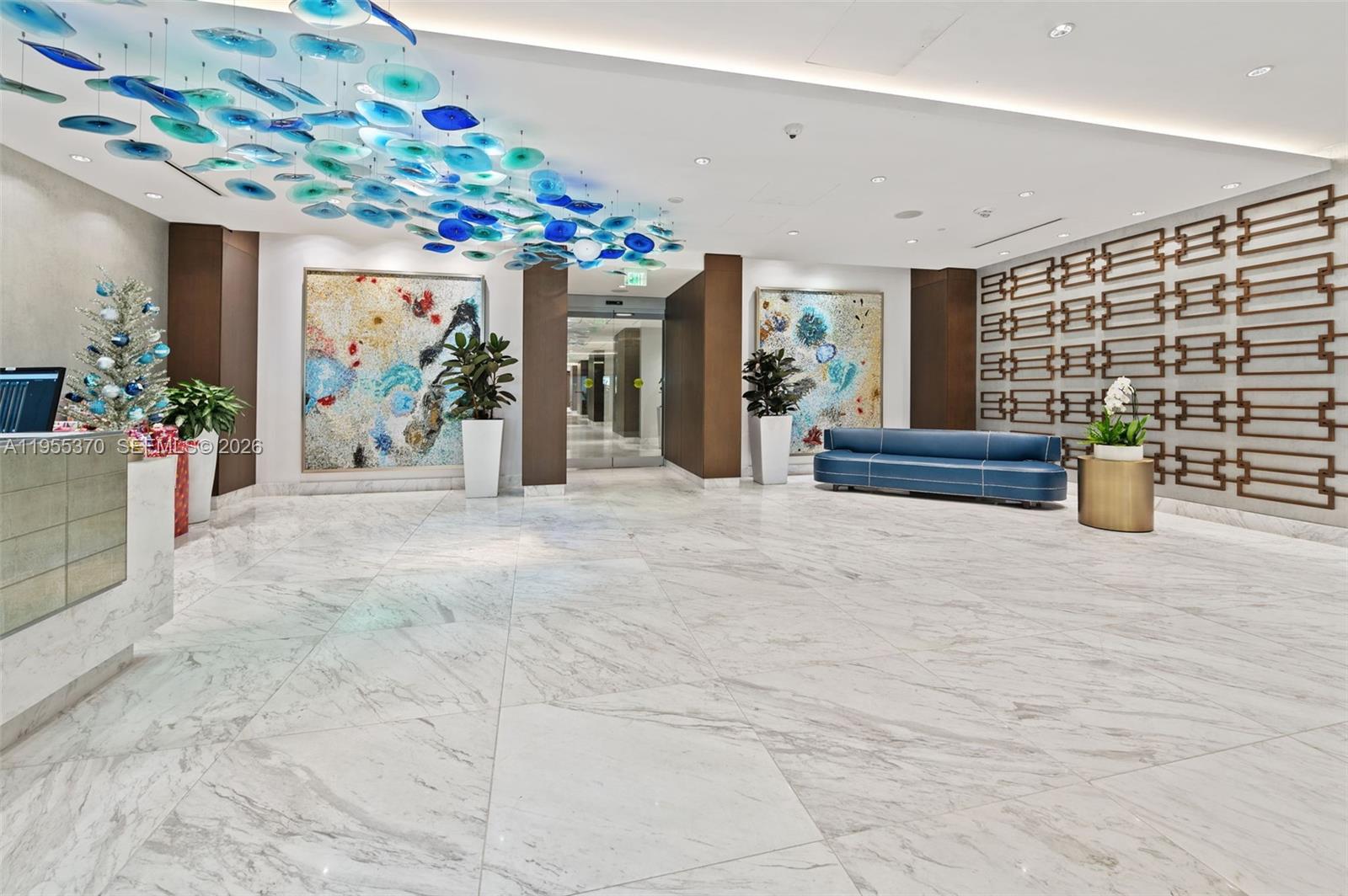 A modern lobby features marble floors, blue glass ceiling art, and a blue sofa — 10155 Collins Ave # 909, Bal Harbour FL 33154, BAL HARBOUR, FL