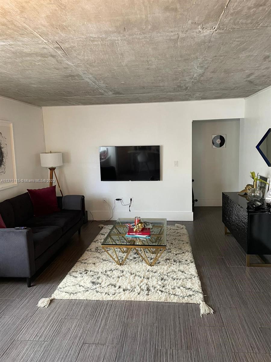 A living room features a dark sofa, glass coffee table, wall-mounted television, and exposed concrete ceiling — 753 Michigan Ave # 2E, Miami Beach FL 33139, MIAMI BEACH, FL