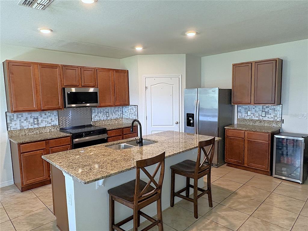 A kitchen features wood cabinets, granite countertops, a central island with seating, and stainless appliances — 1116 FOX PATH ROAD, DAVENPORT, FL
