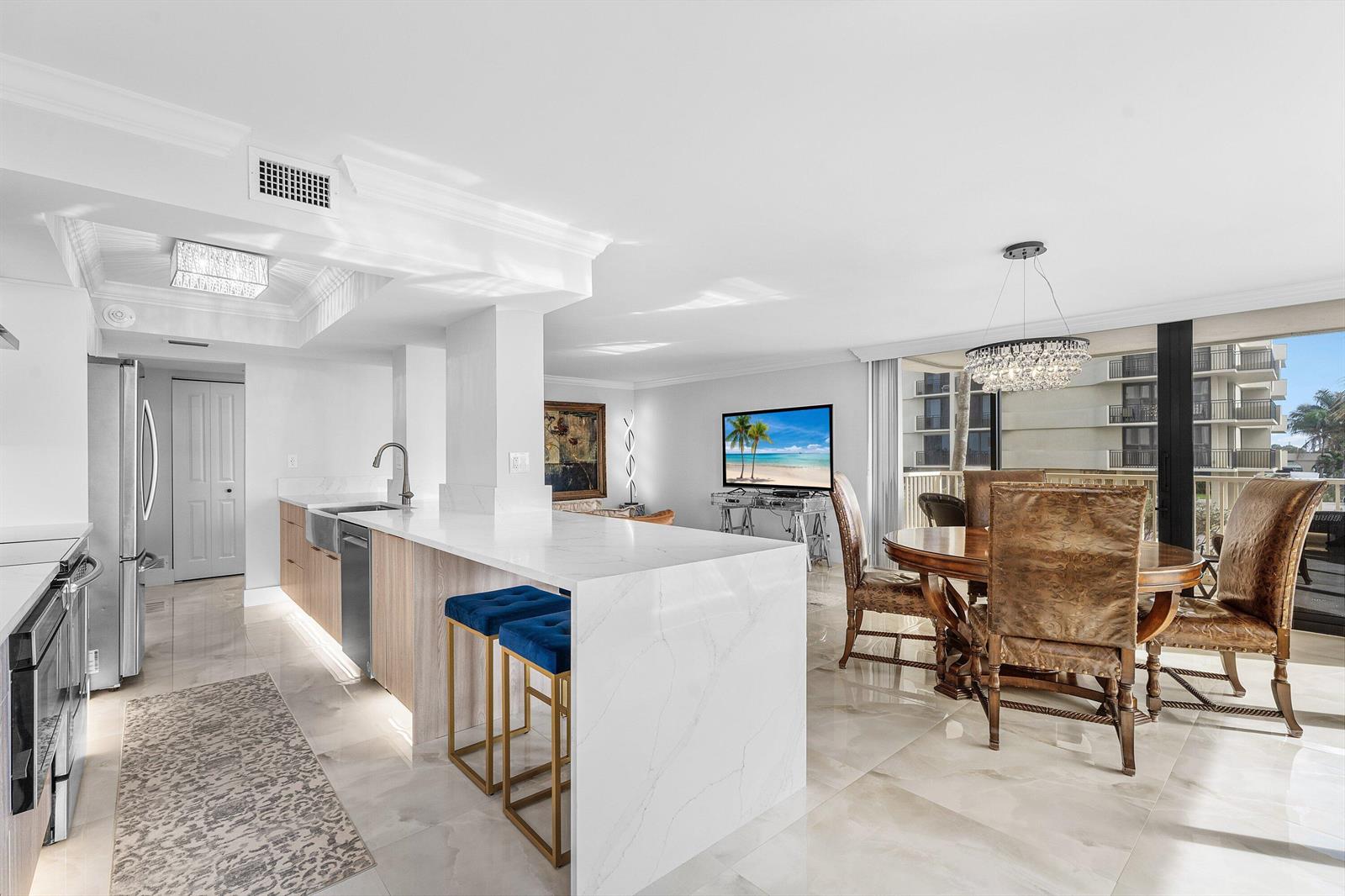 A modern kitchen with a marble waterfall island, blue bar stools, and an adjacent dining area — 5480 N Ocean Drive, Singer Island FL 33404, SINGER ISLAND, FL