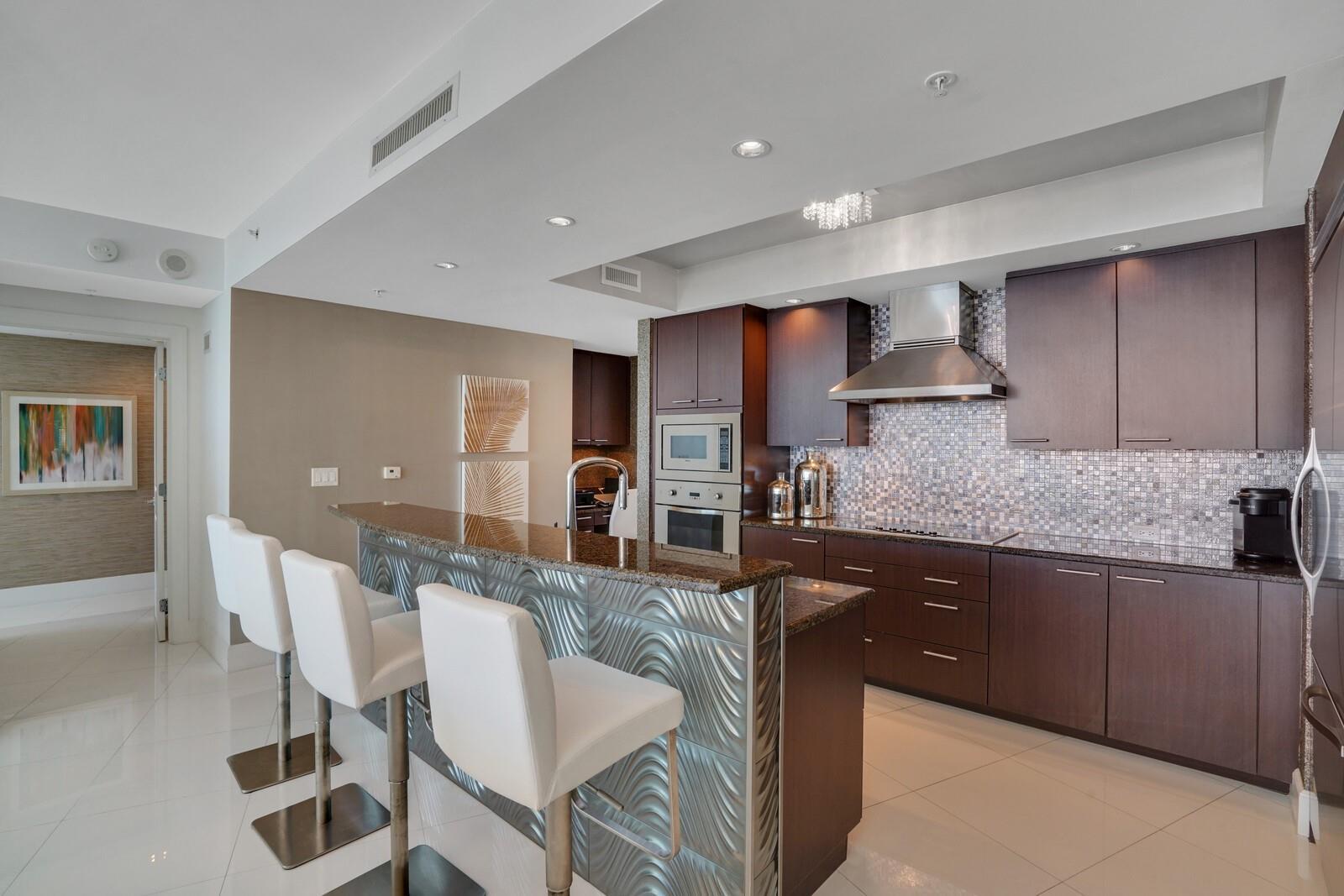 A modern kitchen features dark wood cabinetry, a granite breakfast bar, and white bar stools — 1 N Ocean Boulevard # 903, Pompano Beach FL 33062, POMPANO BEACH, FL