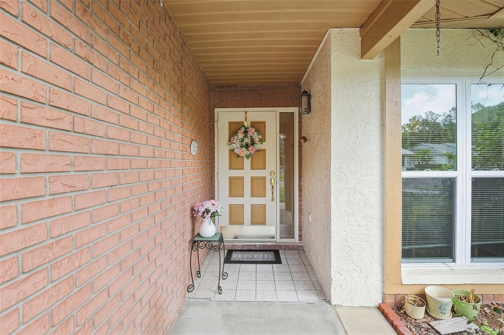 A covered entryway features a brick wall, a white front door with a wreath, and tile flooring — 11622 COCOWOOD DRIVE, NEW PORT RICHEY, FL