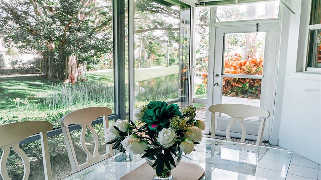 A glass-topped dining table with a floral centerpiece sits inside a sunroom overlooking a yard — 1269 SIESTA BAYSIDE DRIVE UNIT 1269-C, SARASOTA, FL