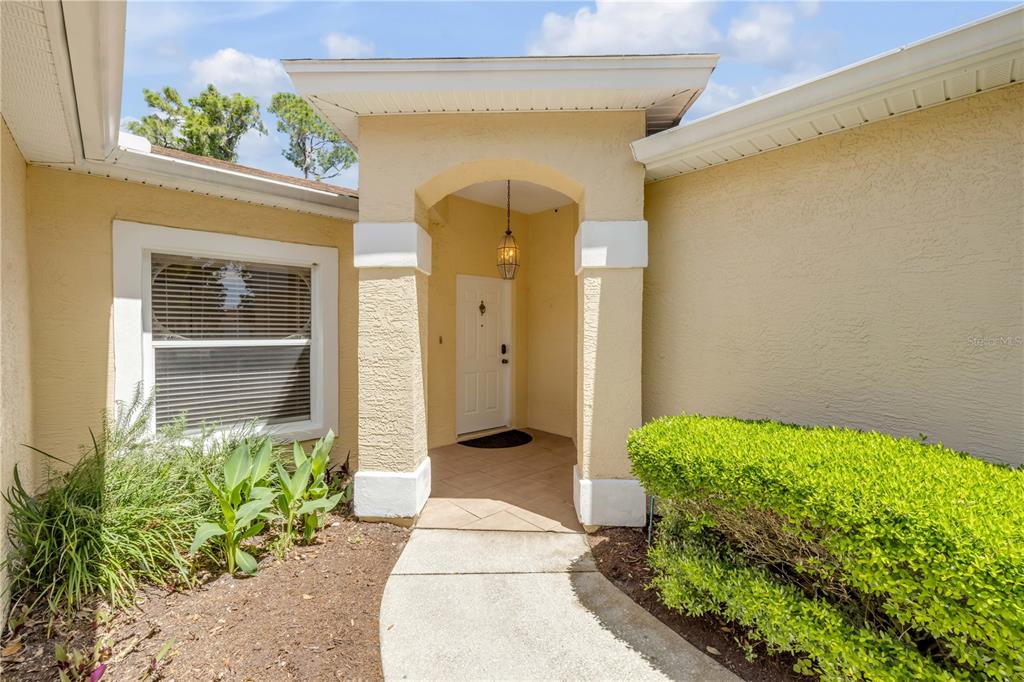 A beige house exterior features a covered entryway, a white front door, and landscaping — 132 TURNBULL VILLAS CIRCLE, NEW SMYRNA BEACH, FL