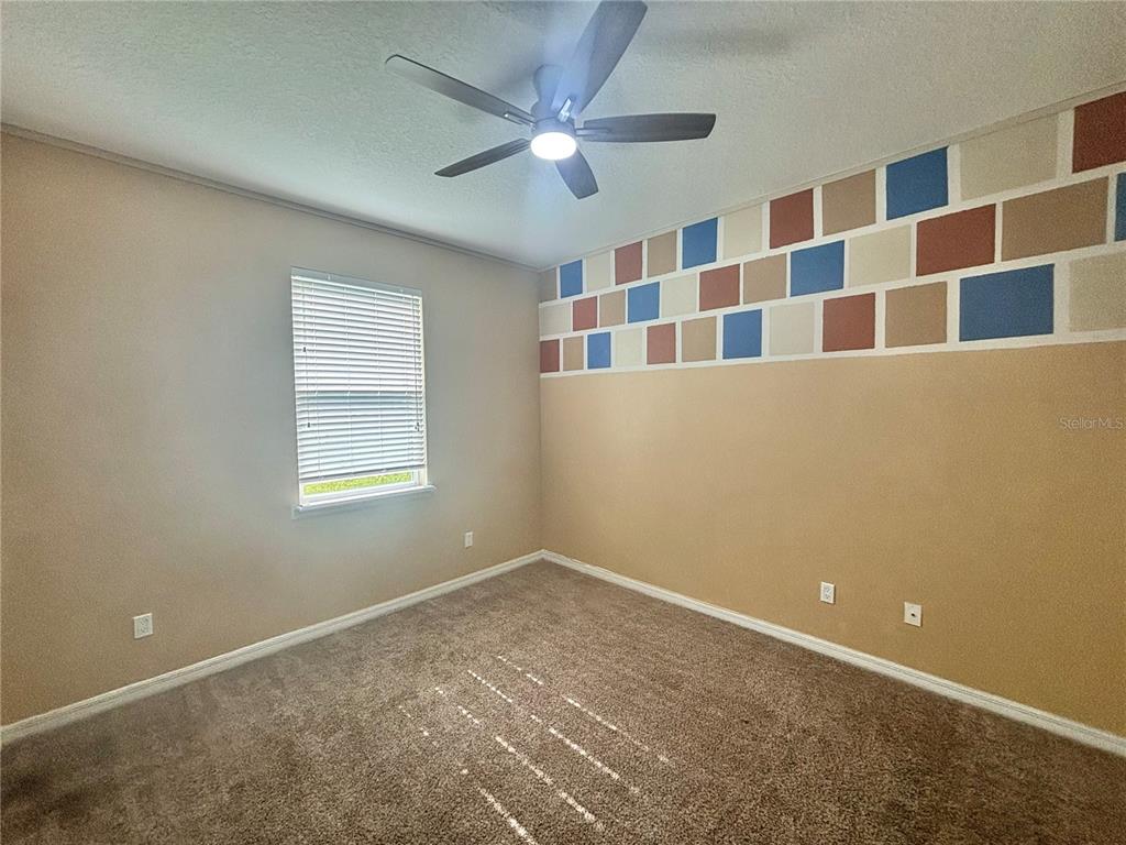 A bedroom with brown carpet, tan walls, a ceiling fan, and a geometric accent wall — 3795 MARIETTA WAY, ST CLOUD, FL