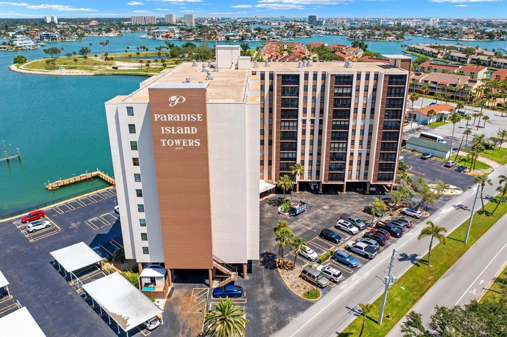 An aerial view shows a multi-story apartment building with a parking lot and waterfront — 10355 PARADISE BOULEVARD UNIT 812, TREASURE ISLAND, FL
