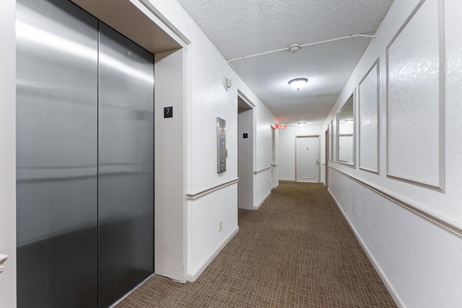 A carpeted apartment hallway features a stainless steel elevator, white walls, and an exit sign — 2425 Presidential Way # 704, West Palm Beach FL 33401, WEST PALM BEACH, FL