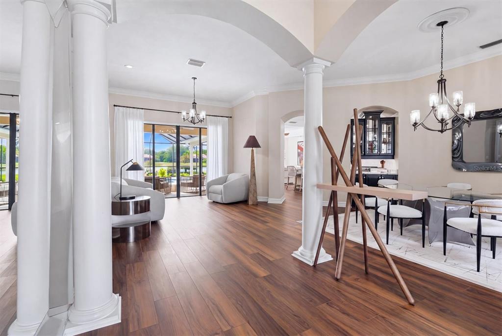 An open-concept living and dining area features white columns, dark wood flooring, and large windows — 6915 HONEYSUCKLE TRAIL, LAKEWOOD RANCH, FL