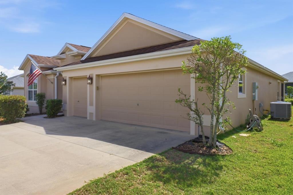 A beige house exterior features a three-car garage, a concrete driveway, and a small tree — 3087 BLACKSTOCK WAY, THE VILLAGES, FL