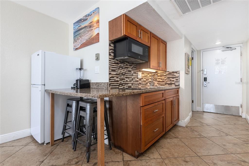 A small kitchen features wood cabinets, granite countertops, a tiled backsplash, and two metal stools — 2301 S ATLANTIC AVENUE UNIT 403, DAYTONA BEACH, FL