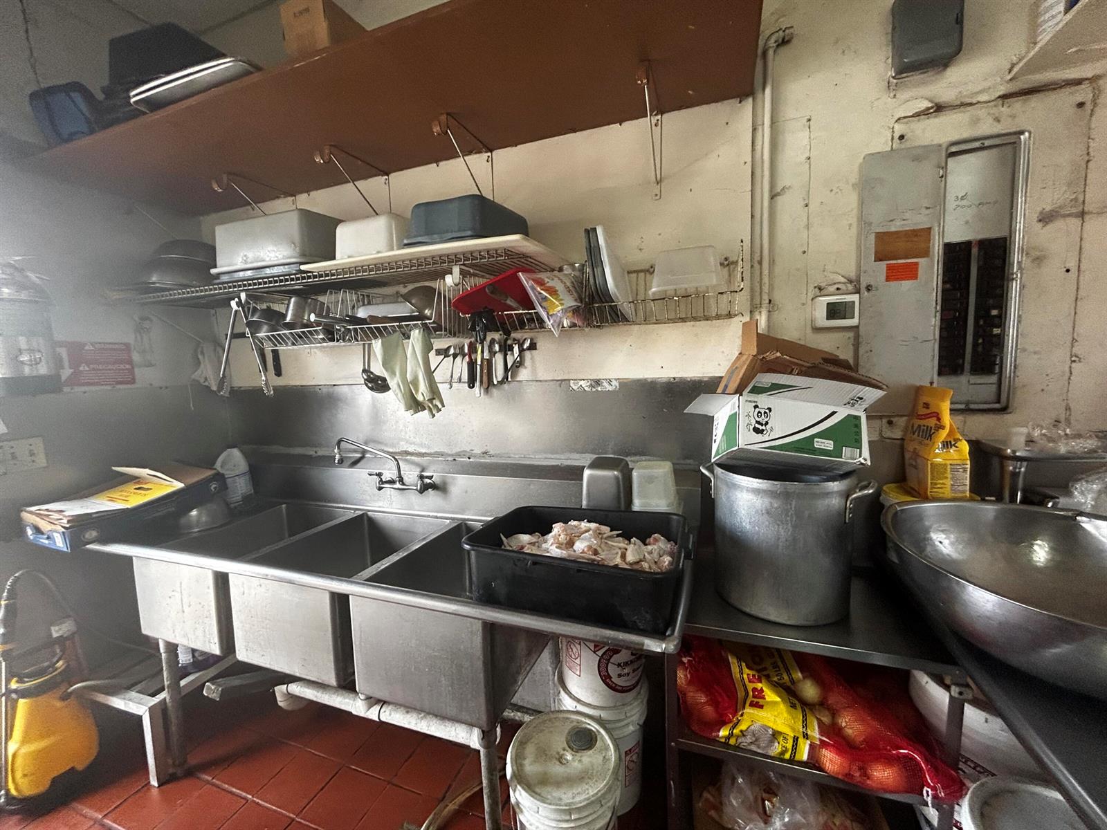 A commercial kitchen features a stainless steel triple-basin sink, metal shelving, and food storage containers — 2623 W Atlantic Boulevard, Pompano Beach FL 33069, POMPANO BEACH, FL