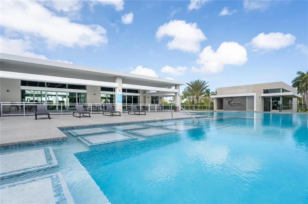 A swimming pool with a sun shelf sits in front of a modern clubhouse building — Calle Central Park URB. RIVIERA VILLAGE, BAYAMON, PR