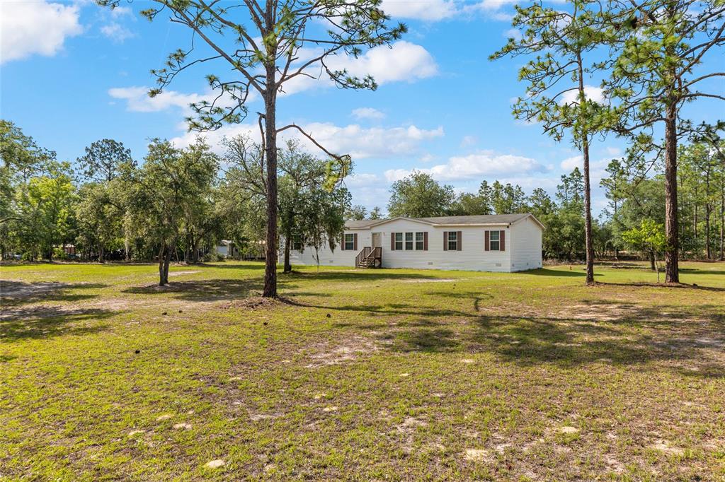 A single-story manufactured home sits on a large, grassy lot surrounded by pine trees — 14671 SE 5TH PLACE, WILLISTON, FL
