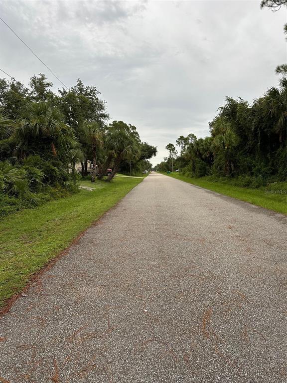 A paved road stretches into the distance, flanked by grassy shoulders and dense green trees — 483 TROUTDALE STREET, PORT CHARLOTTE, FL