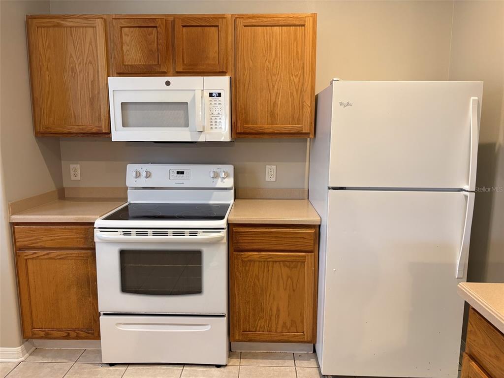 A kitchen features oak cabinets, white appliances, and tan countertops against a neutral wall — 3922 WASHBURN PLACE, WESLEY CHAPEL, FL