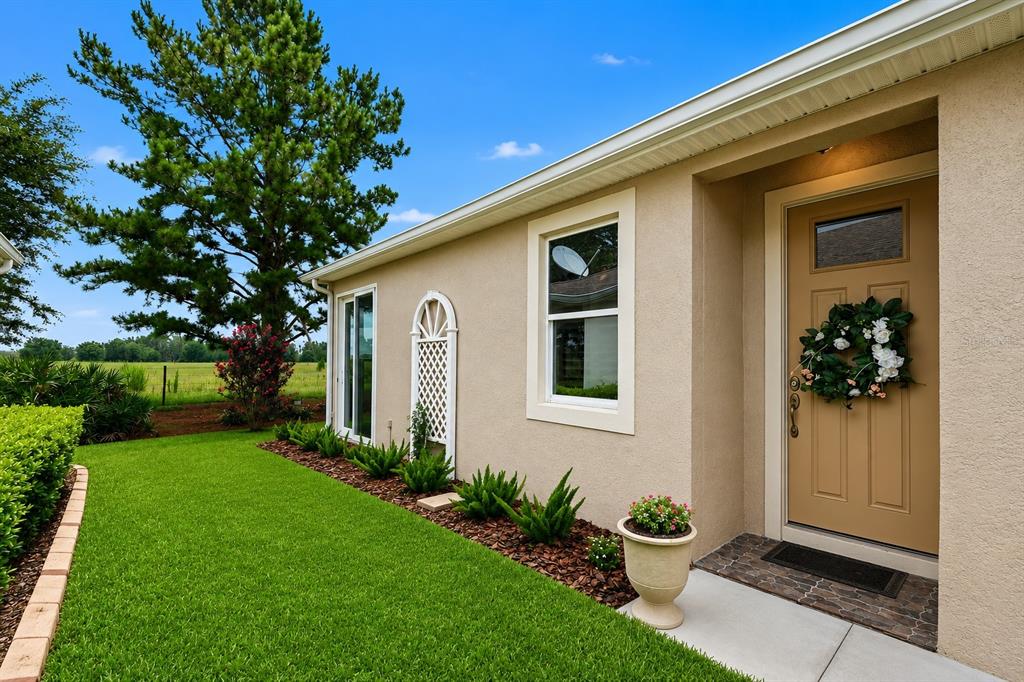 A tan house exterior features a front door with a wreath, a window, and landscaping — 32983 WINDELSTRAW DRIVE, WESLEY CHAPEL, FL