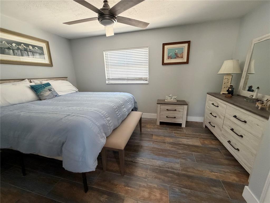A bedroom features a bed with a blue comforter, wood-look tile flooring, and white furniture — 5053 N BEACH ROAD UNIT 9E/W, ENGLEWOOD, FL