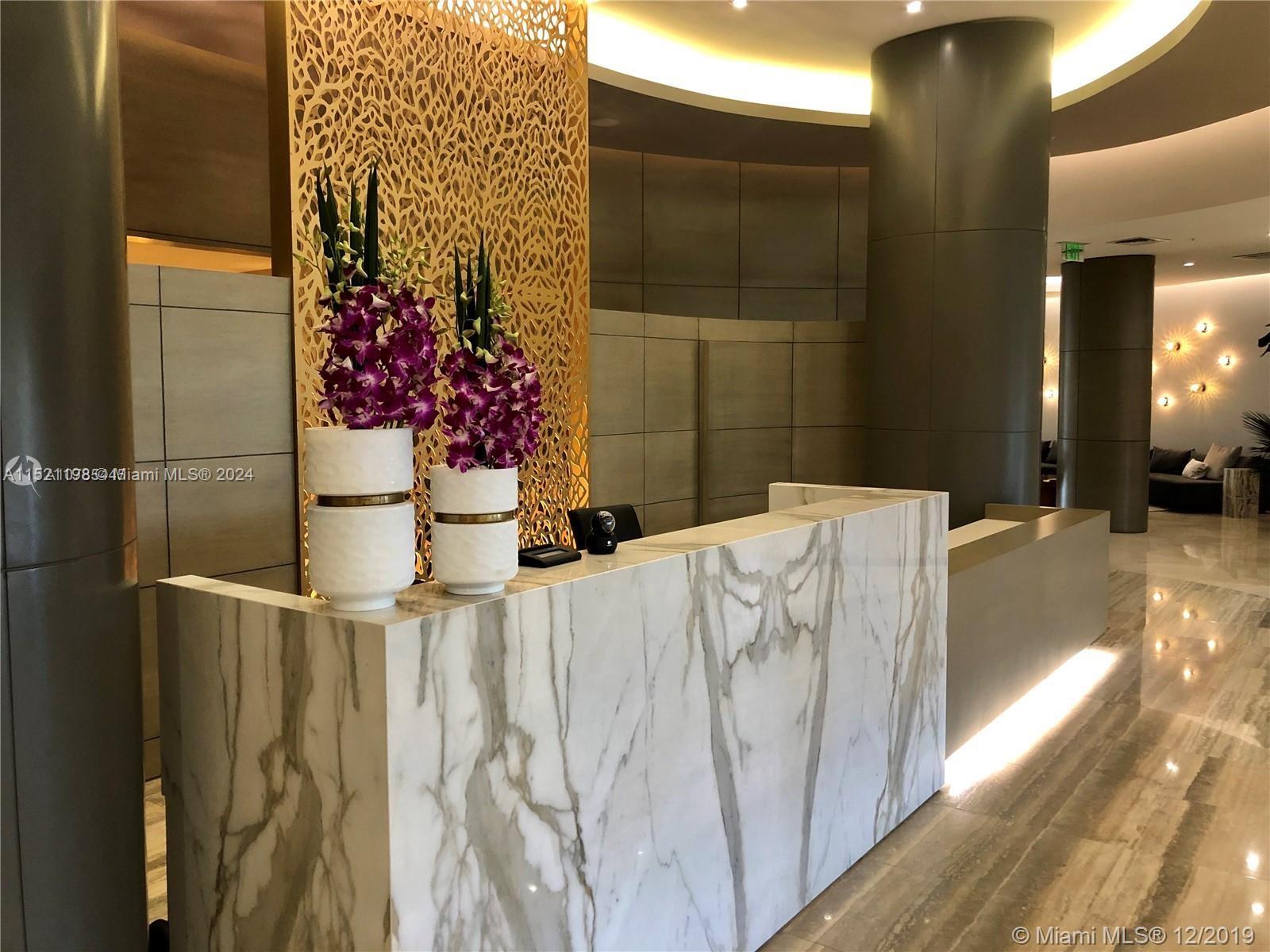 A modern lobby features a marble reception desk, decorative gold screen, and two floral arrangements — 10275 Collins Ave # 230, Bal Harbour FL 33154, BAL HARBOUR, FL