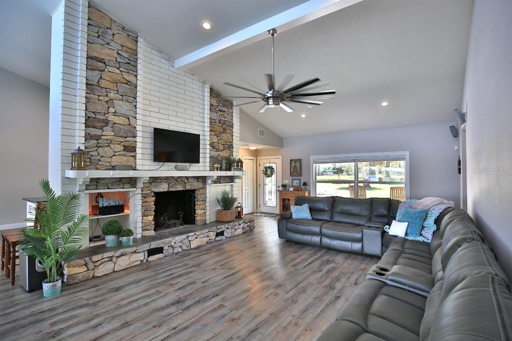 A living room features a large stone fireplace, vaulted ceiling, ceiling fan, and gray flooring — 10893 SE 141 AVENUE ROAD, OCKLAWAHA, FL