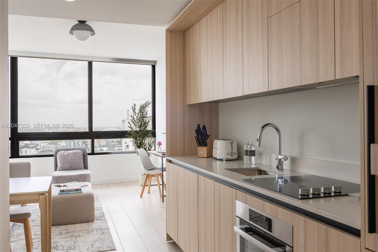 A modern kitchen with light wood cabinetry overlooks a living area with floor-to-ceiling windows — 225 N Miami Ave # 3412, Miami FL 33128, MIAMI, FL