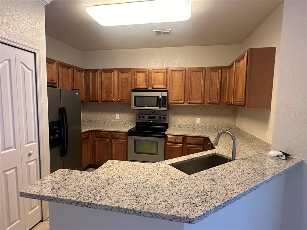 A kitchen features wood cabinets, granite countertops, stainless steel appliances, and a peninsula sink — 6119 METROWEST BOULEVARD UNIT 103, ORLANDO, FL