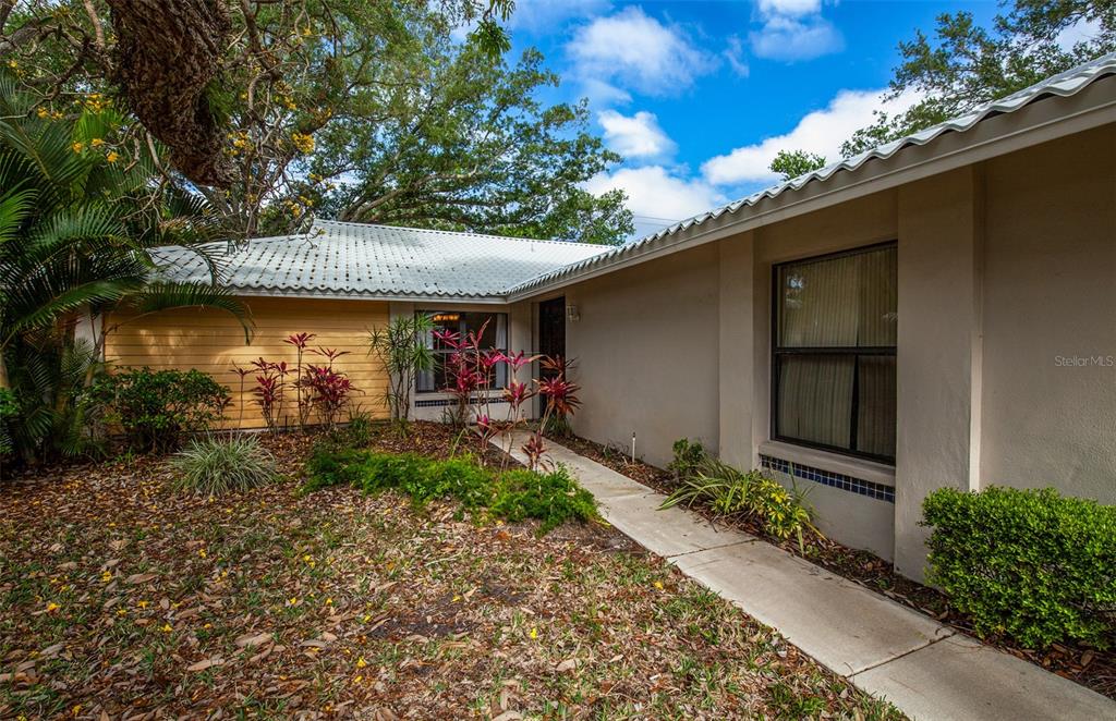 The exterior of a house features a light-colored stucco wall, a metal roof, and landscaping — 247 SOUTHAMPTON LANE UNIT 272, VENICE, FL