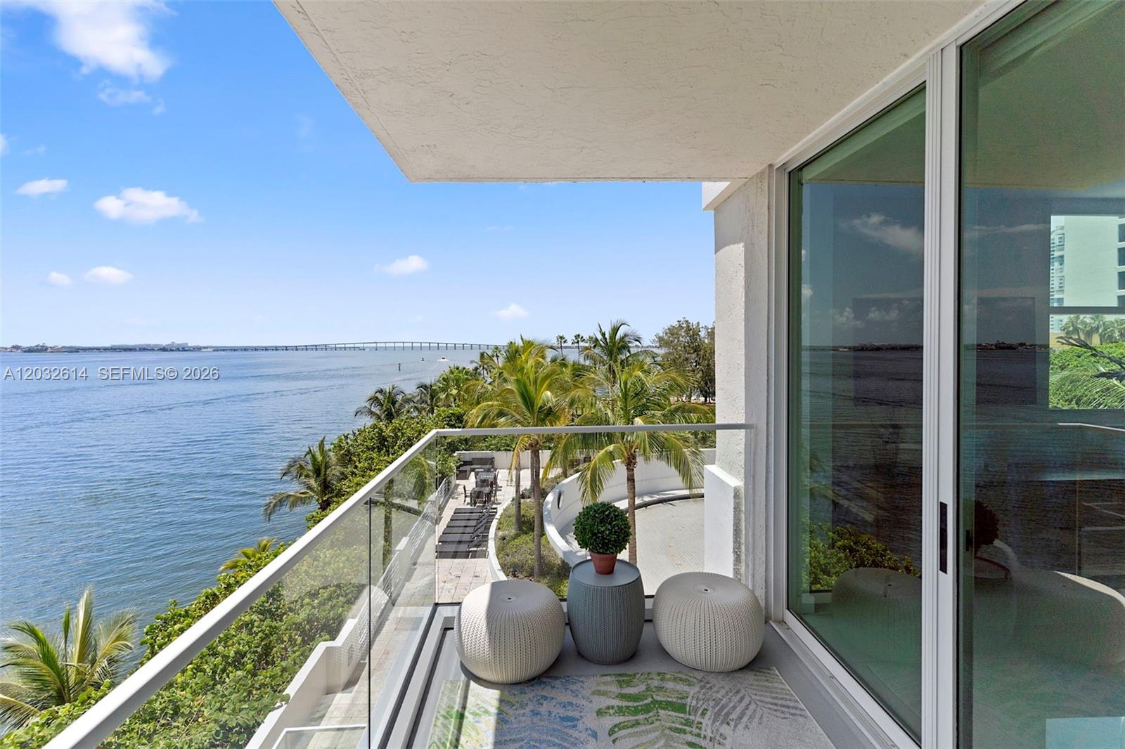 A balcony features a glass railing, two white ottomans, a small table, and water views — 800 CLAUGHTON ISLAND DRIVE # 505, Miami FL 33131, MIAMI, FL