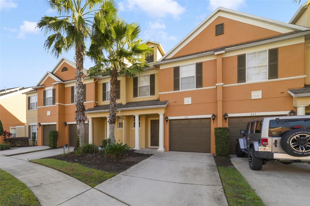 A row of two-story townhomes with orange stucco facades, brown garage doors, and palm trees — 851 ASSEMBLY COURT, KISSIMMEE, FL