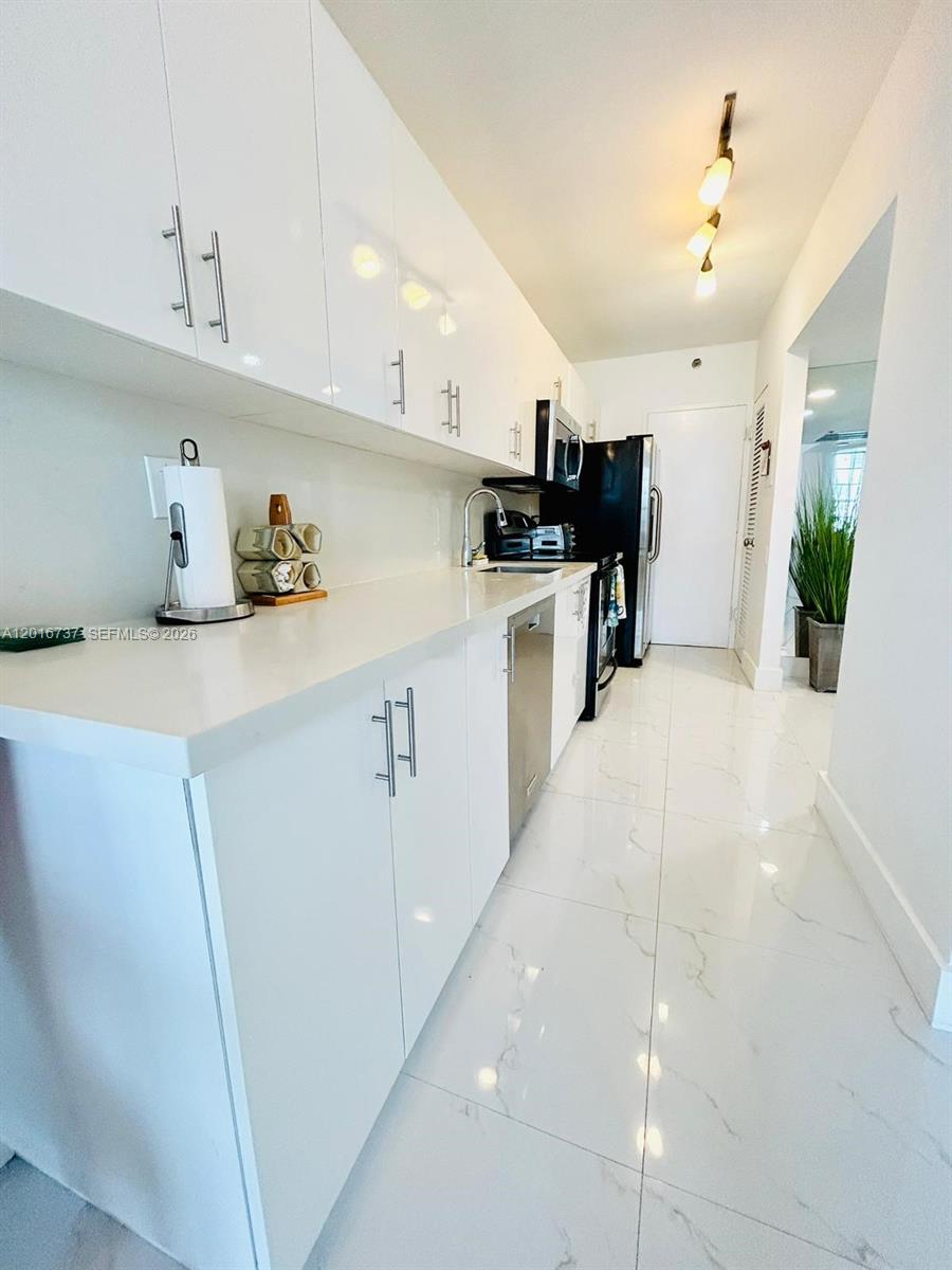 A galley kitchen features white cabinets, stainless steel appliances, and white marble-look tile flooring — 100 Lincoln Rd # 1526, Miami Beach FL 33139, MIAMI BEACH, FL