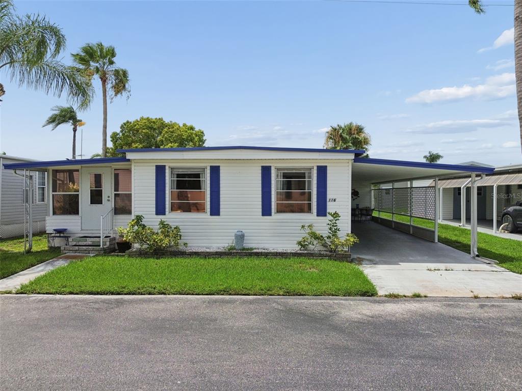 A white mobile home with blue shutters and a carport sits behind a grassy lawn — 18675 US HIGHWAY 19 N UNIT 116, CLEARWATER, FL