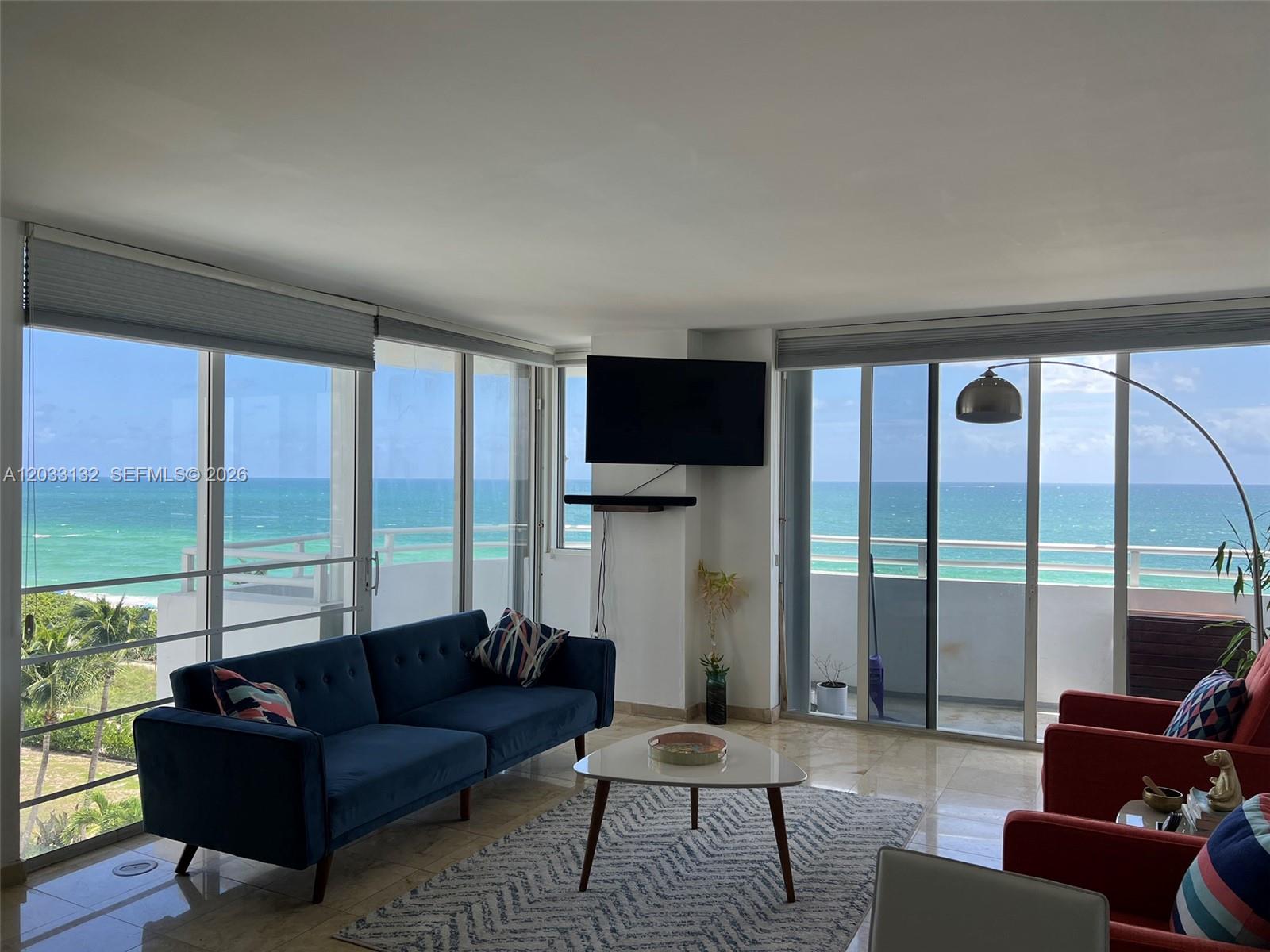 A living room features a blue sofa, red armchairs, and floor-to-ceiling windows overlooking the ocean — 8911 Collins Ave # 605, Surfside FL 33154, SURFSIDE, FL