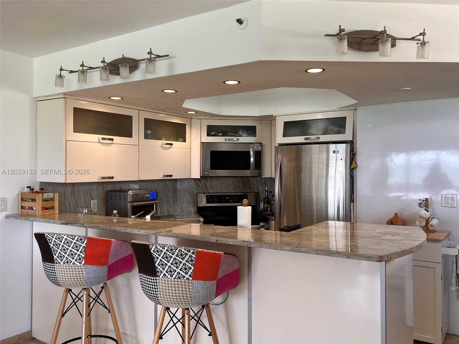 A kitchen features white cabinets, stainless steel appliances, granite countertops, and two patterned bar stools — 8911 Collins Ave # 605, Surfside FL 33154, SURFSIDE, FL