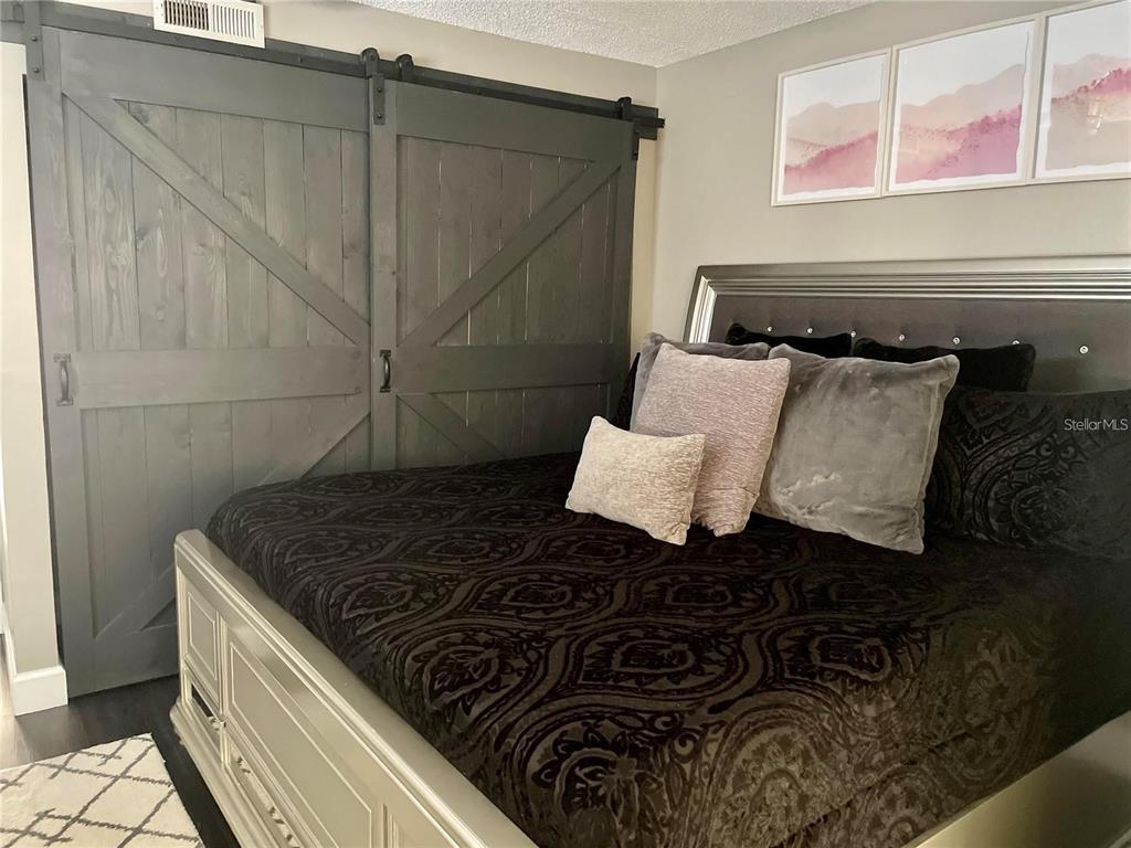 A bedroom features a dark wood barn door, a bed with patterned bedding, and wall art — 2571 CYPRUS DRIVE UNIT 1-204, PALM HARBOR, FL