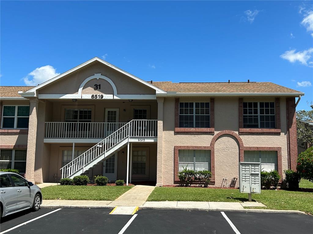 A two-story apartment building features a tan stucco exterior, brick accents, and an outdoor staircase — 6519 SPRING FLOWER DRIVE UNIT 24, NEW PORT RICHEY, FL