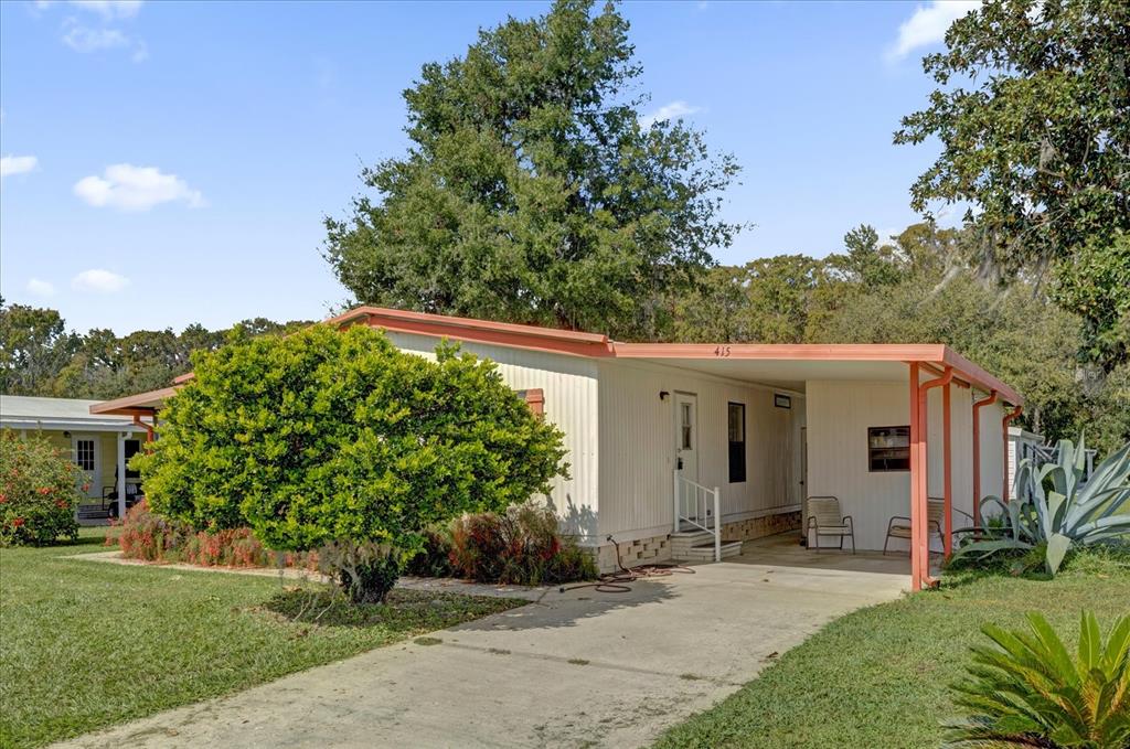 A single-story mobile home with a carport, white siding, and a large front tree — 415 S TIMBER TRAIL, WILDWOOD, FL