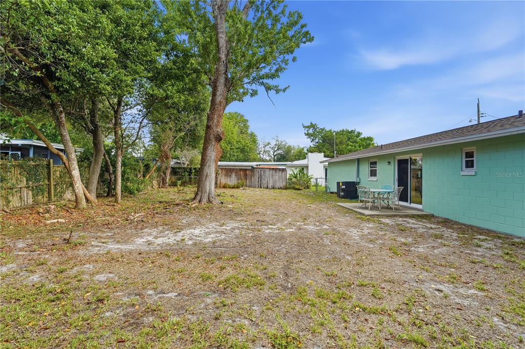 A backyard with a sandy lawn, large trees, a wooden fence, and a patio area — 1528 VALENCIA AVENUE, DAYTONA BEACH, FL