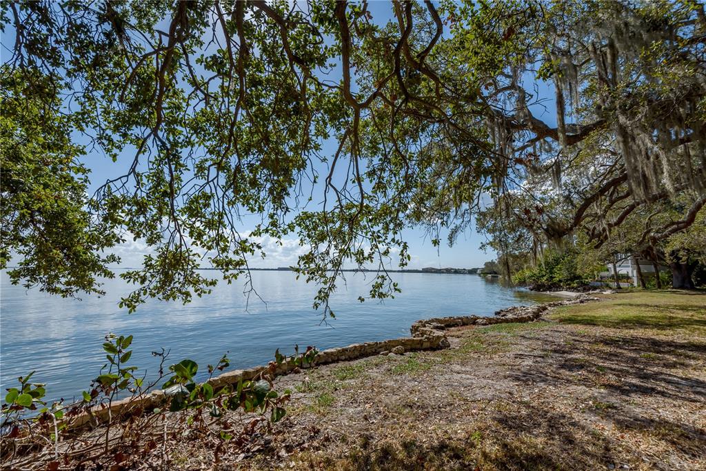 A stone-lined shoreline overlooks a calm body of water beneath large, moss-draped oak trees — 6210 BAYSHORE BOULEVARD, TAMPA, FL
