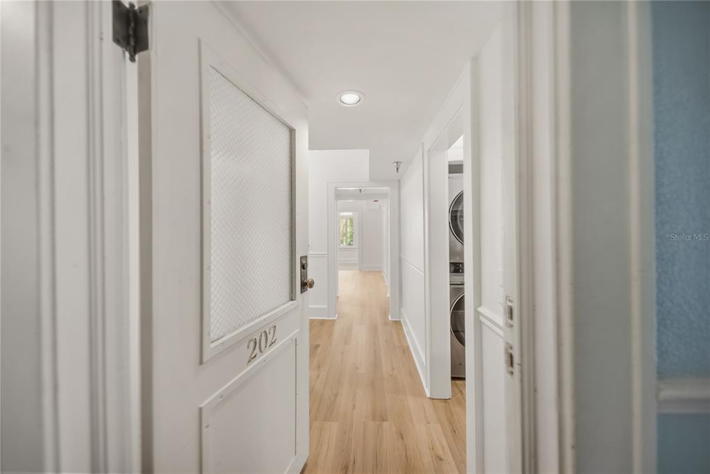 A hallway with light wood flooring leads past a laundry closet to a distant window — 1221 N PALM AVENUE UNIT 202, SARASOTA, FL