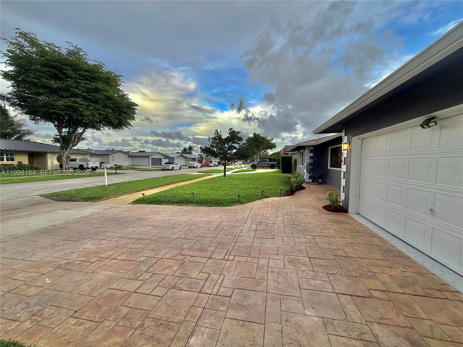 A paved driveway leads to a garage next to a lawn with trees under clouds — 8404 SW 26th St, Davie FL 33324, DAVIE, FL
