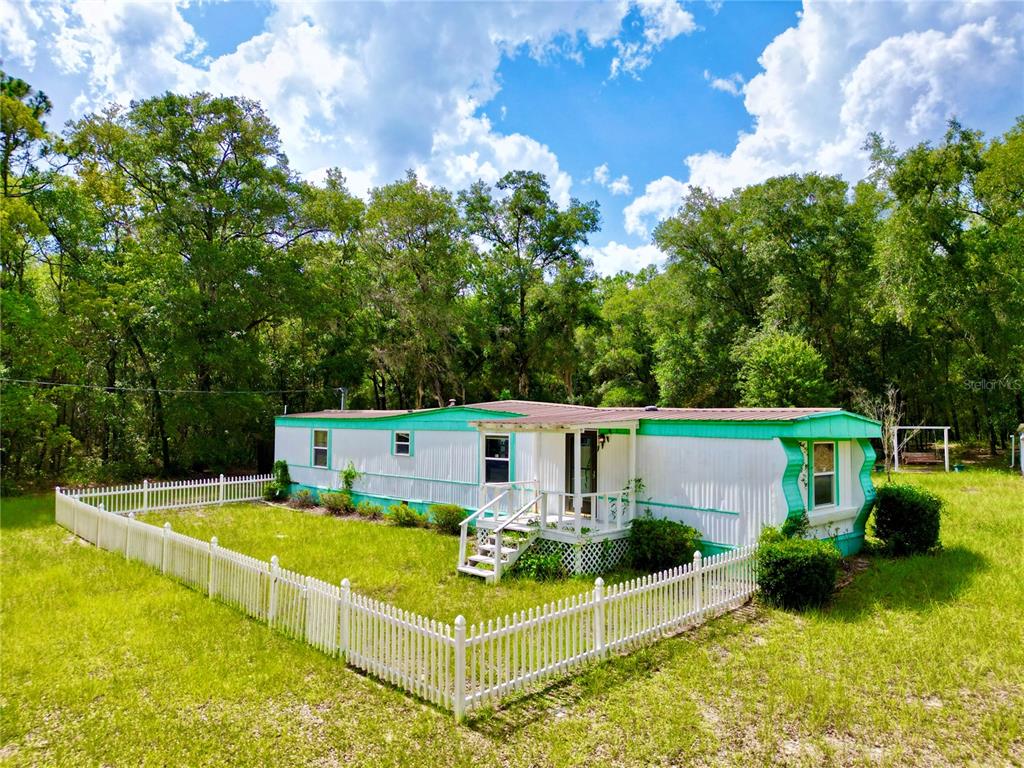 A white mobile home with teal trim sits behind a white picket fence near woods — 8232 SWAN LAKE ROAD, MELROSE, FL