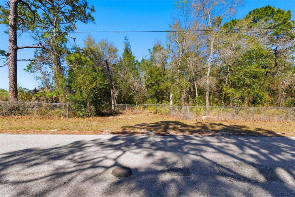 A vacant, fenced-in residential lot sits alongside a paved road under a clear blue sky — 9255 GROUSE WAY, HUDSON, FL