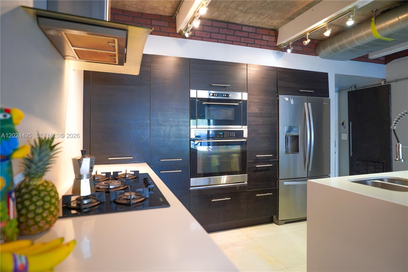 A modern kitchen features dark wood cabinetry, stainless steel appliances, and an exposed brick wall — 18101 NW 68th Ave # C204, Hialeah FL 33015, HIALEAH, FL