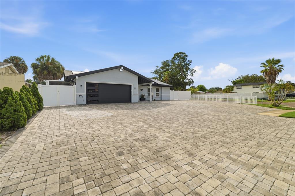 A house exterior features a large paver driveway, a black garage door, and white fencing — 7101 SHENANDOAH COURT, TAMPA, FL