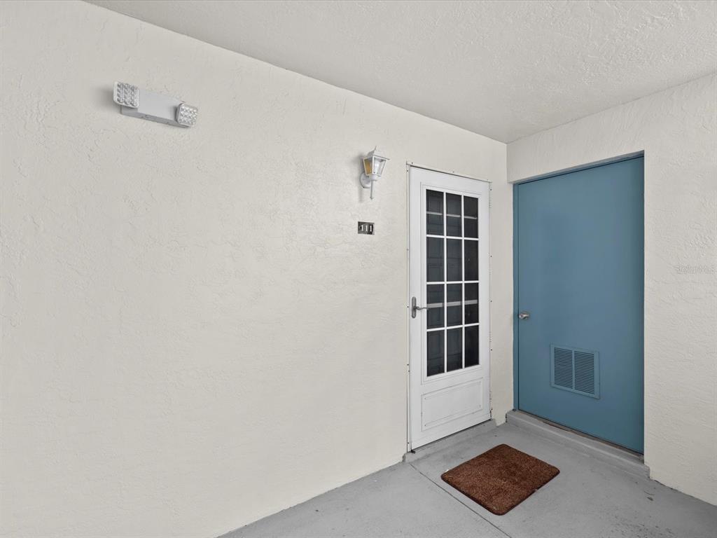 A white exterior wall features a blue door, a storm door, and unit number 111 — 2737 ENTERPRISE ROAD E UNIT 111, CLEARWATER, FL