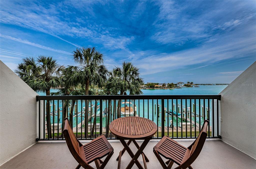 A balcony features a small wooden table and two chairs overlooking a tropical waterfront view — 9495 BLIND PASS ROAD UNIT 205, ST PETE BEACH, FL