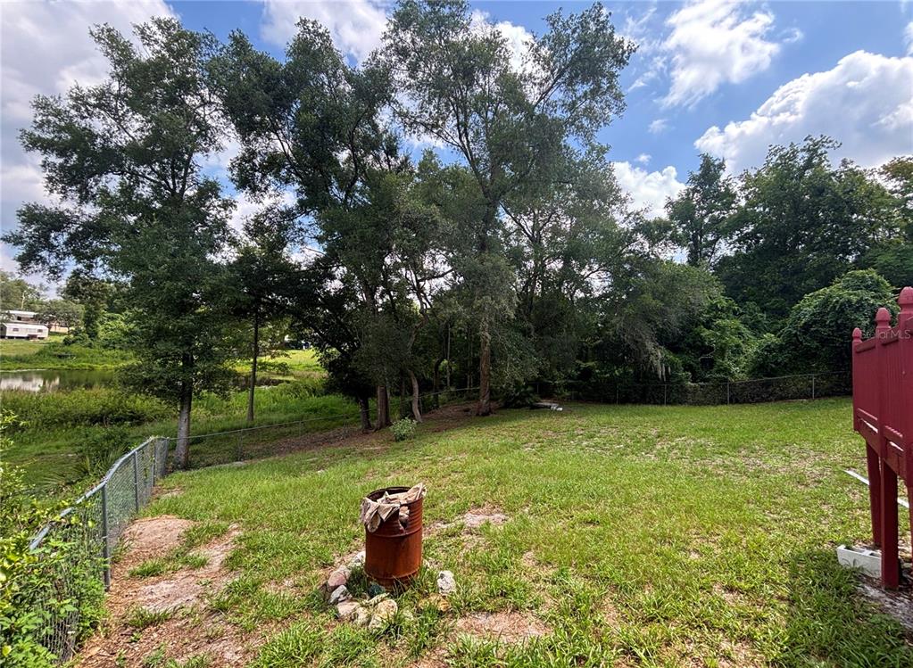 A grassy backyard features a rusty metal barrel, mature trees, and a chain-link fence — 18225 SE 22ND PLACE, SILVER SPRINGS, FL