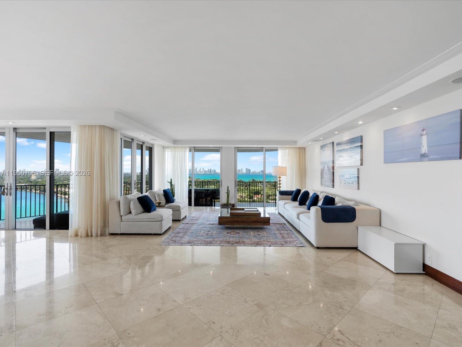 A spacious living room features light tile floors, white sofas, and floor-to-ceiling windows overlooking water — 5660 Collins Ave # 18A, Miami Beach FL 33140, MIAMI BEACH, FL