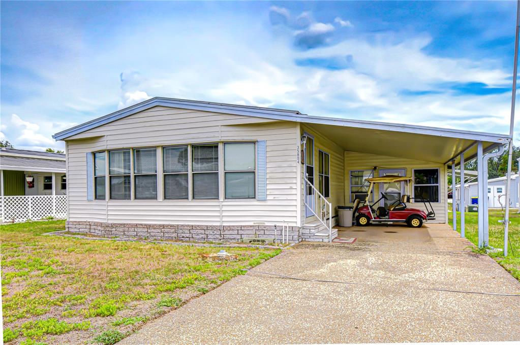 A single-story manufactured home with light siding, a stone-patterned base, and an attached carport — 35804 BRISA DRIVE, ZEPHYRHILLS, FL