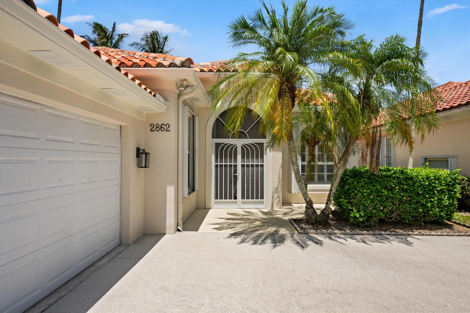A beige house exterior features a white garage door, arched entryway, and palm trees — 2862 Irma Lake Drive, West Palm Beach FL 33411, WEST PALM BEACH, FL